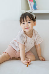 Toddler girl smiling in Pure Hug rosy blush summer set, sitting on a sofa, wearing pink shorts and floral top.